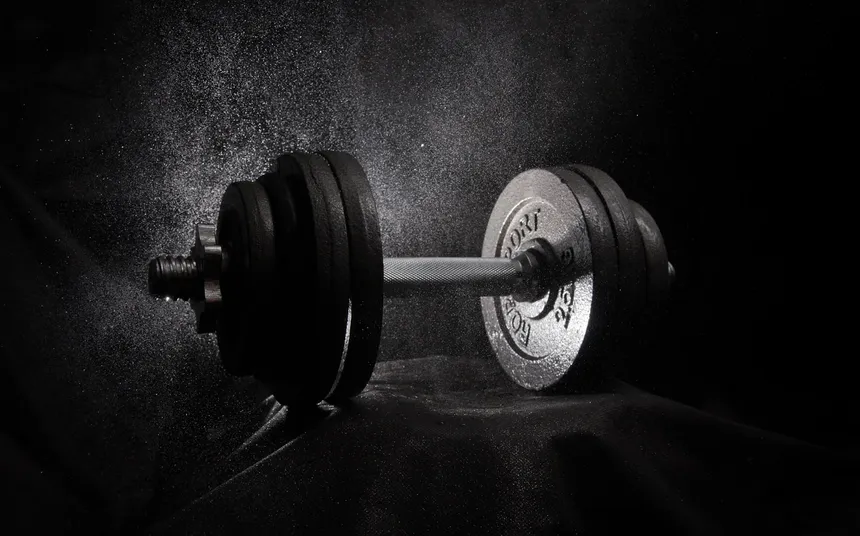A single adjustable dumbbell lit from one side against a dark background, dust hanging in the air around it