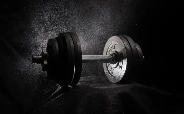 A single adjustable dumbbell lit from one side against a dark background, dust hanging in the air around it
