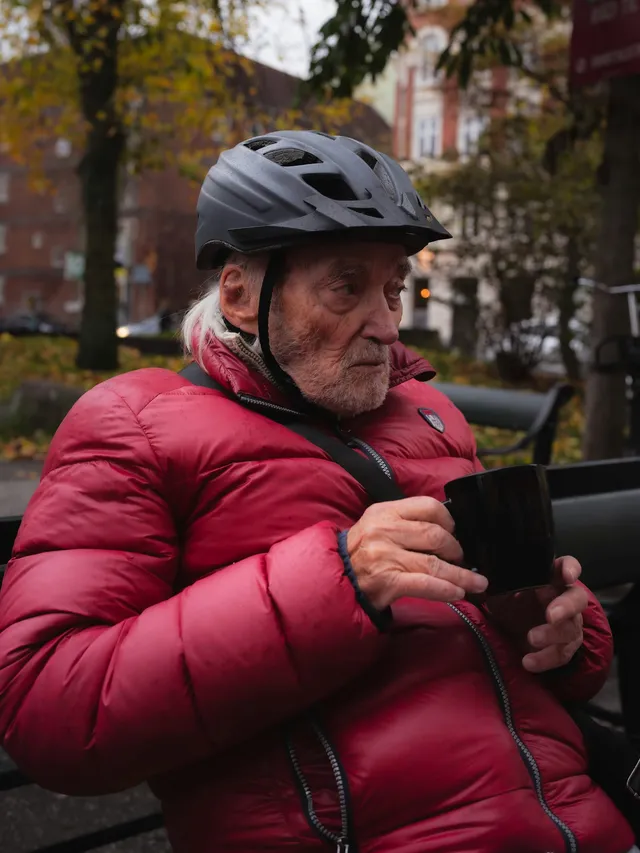 An older man in a warm jacket and a cycling helmet pausing outdoors with a hot drink