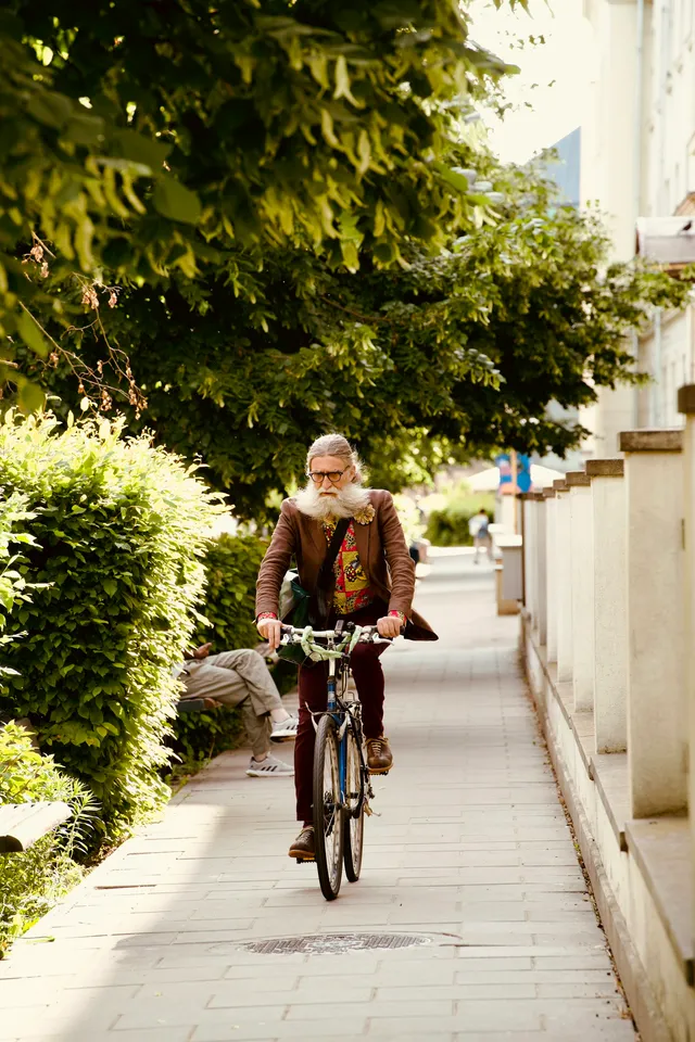 An older man in a jacket and long trousers riding an ordinary two-wheeled bicycle along a tree-lined path