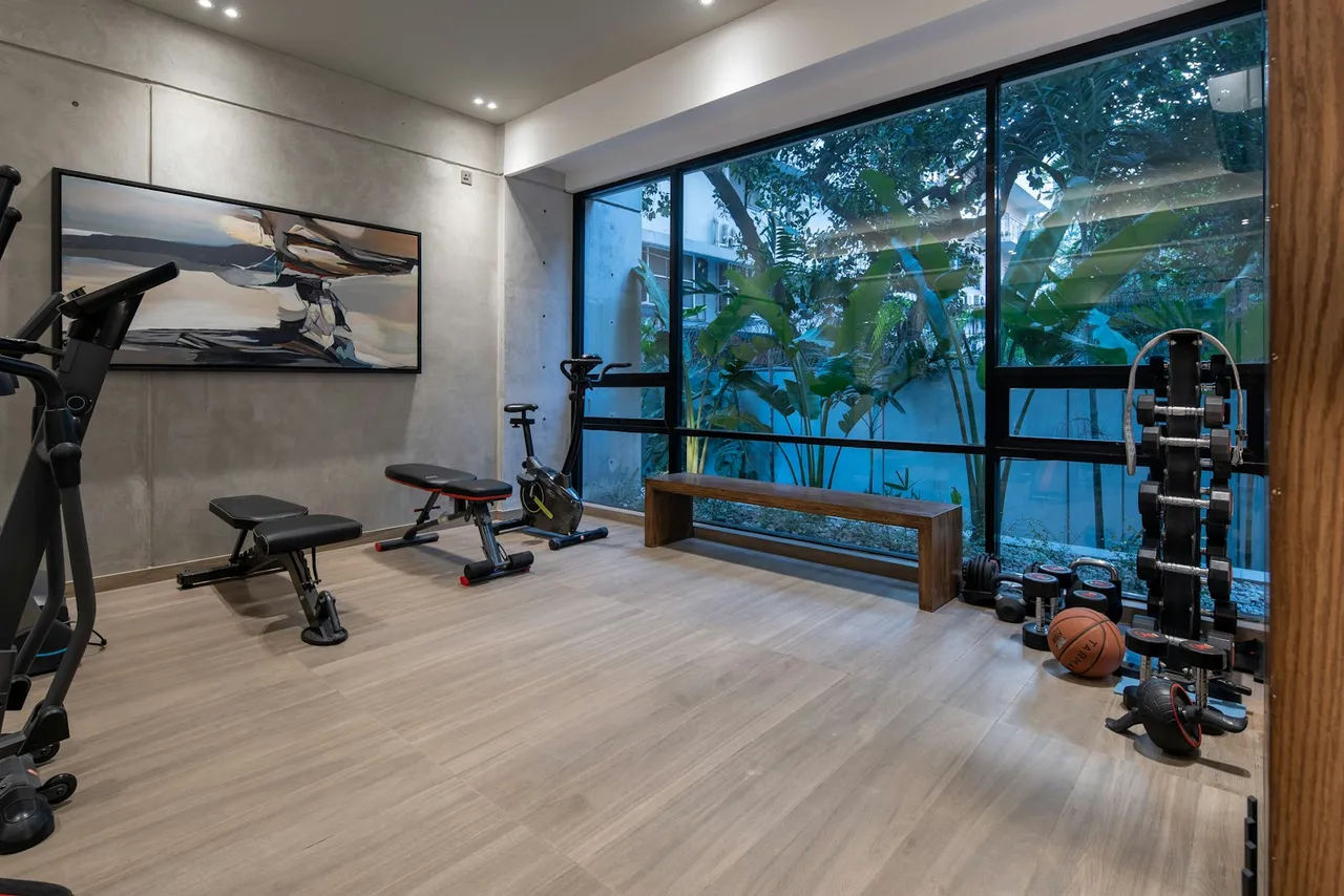 A home gym along one wall of a bright room: a rack of dumbbells, a bench, a cable machine and a mat, with daylight from a full-height window
